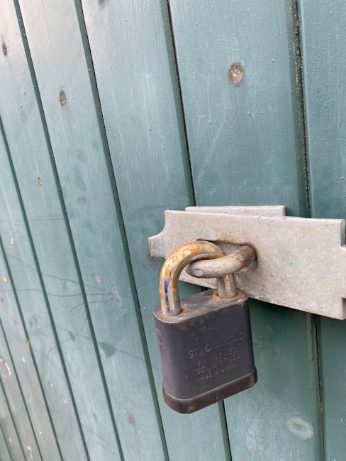 A close-up image of a black padlock securing a metal latch on a vertical green wooden fence or gate. The padlock has a slightly weathered appearance, with some rust visible on the shackle, which is th